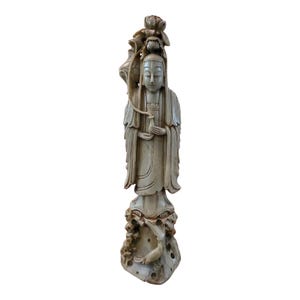 May include: A white stone sculpture of a female figure standing on a base with a carved fish. The figure is wearing a long robe and has a flower crown on her head.