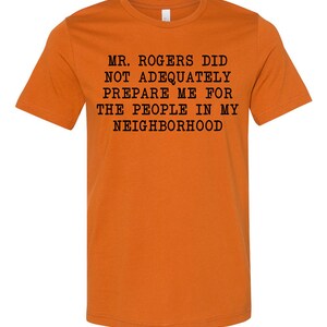 May include: Orange t-shirt with black text that reads "Mr. Rogers did not adequately prepare me for the people in my neighborhood."