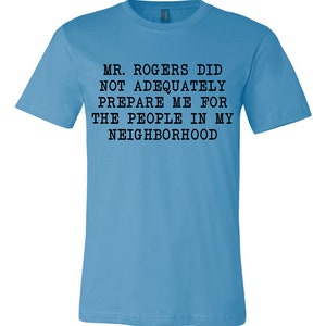 May include: A light blue t-shirt with black text that reads "MR. ROGERS DID NOT ADEQUATELY PREPARE ME FOR THE PEOPLE IN MY NEIGHBORHOOD".