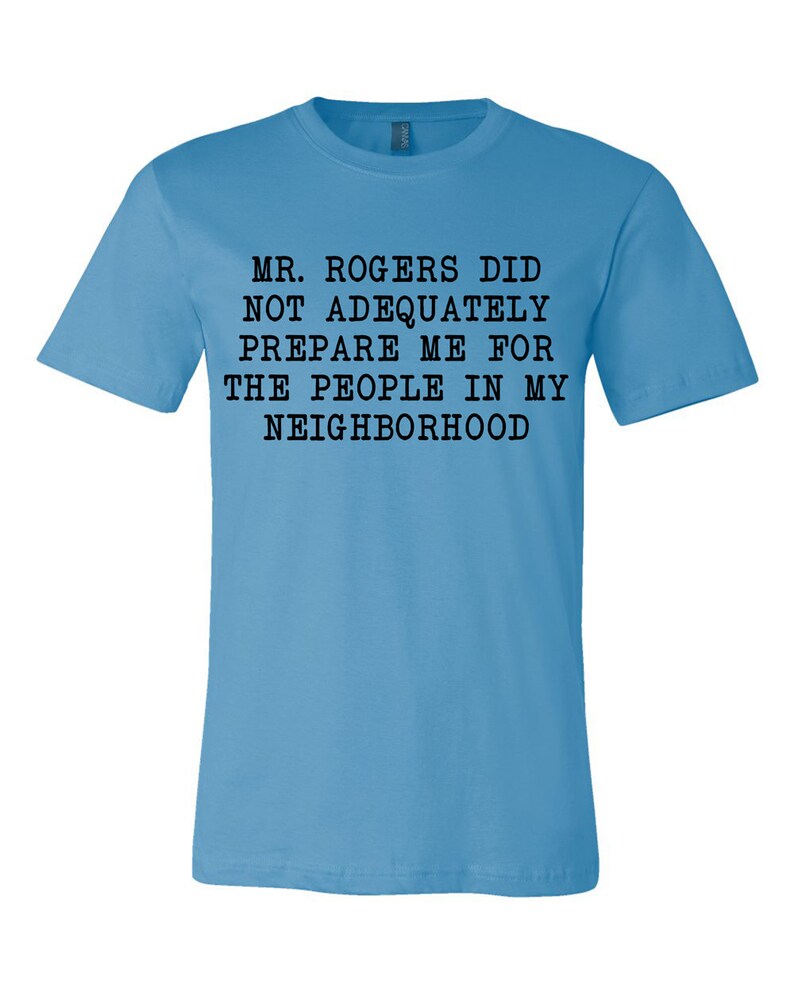 May include: A light blue t-shirt with black text that reads "MR. ROGERS DID NOT ADEQUATELY PREPARE ME FOR THE PEOPLE IN MY NEIGHBORHOOD".