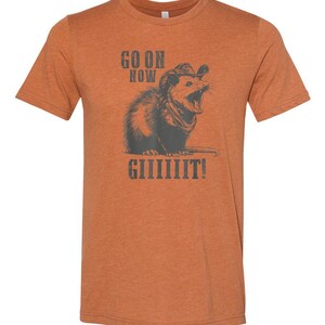 May include: Orange t-shirt with a graphic of an opossum wearing a cowboy hat and bandana, with the text "GO ON NOW GIIIIIIIT!". The opossum is in shades of gray, and the text is in a dark gray color.