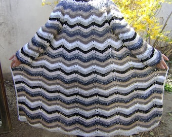 May include: A long, handmade crochet cardigan in a chevron pattern. The color palette includes shades of white, black, gray, and tan. The sleeves are long, and the cardigan is open in the front. The garment is a cozy, handmade piece.