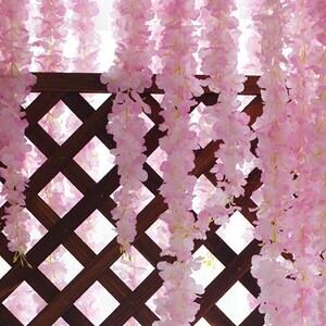 Könnte beinhalten: Rosa künstliche Glyzinienblüten, die vor einem braunen Gitterzaun hängen. Die Blüten sind in langen Strängen angeordnet und erzeugen einen kaskadierenden Effekt.