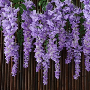 May include: A long strand of artificial wisteria flowers with purple blooms hanging from a brown bamboo background.