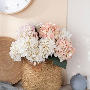 May include: A bouquet of pink and white hydrangeas in a woven basket. The flowers are arranged in a vase and are sitting on a table.