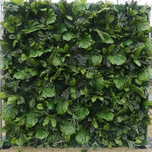 May include: A large, square artificial green wall composed of various types of lush, vibrant green leaves. The leaves include large, heart-shaped monstera leaves, ferns, and other tropical foliage. The wall is mounted on a metal frame and set outdoors.
