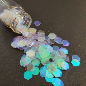 May include: A glass jar with a white lid spills iridescent hexagonal glitter onto a black surface. The glitter is a mix of clear, light blue, and light purple.
