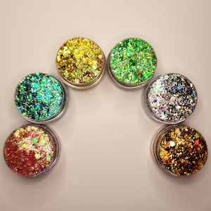 May include: Six small jars of glitter, each containing a different color and texture of glitter. The jars are arranged in a semi-circle on a white surface.