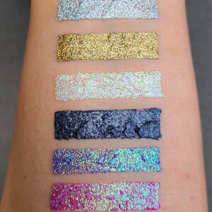K&ouml;nnte beinhalten: Sechs verschiedene Glitzer-Eyeliner-Swatches auf dem Arm einer Person. Die Swatches sind silber, gold, schimmernd wei&szlig;, schwarz, blau und pink.