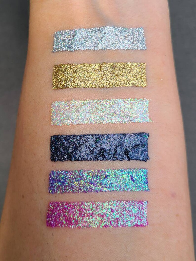 K&ouml;nnte beinhalten: Sechs verschiedene Glitzer-Eyeliner-Swatches auf dem Arm einer Person. Die Swatches sind silber, gold, schimmernd wei&szlig;, schwarz, blau und pink.