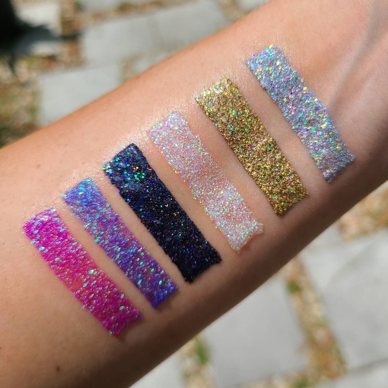 K&ouml;nnte beinhalten: Sechs verschiedene Farbt&ouml;ne von Glitzer-Eyeliner-Swatches auf dem Arm einer Person. Die Farben sind rosa, lila, blau, schwarz, wei&szlig; und gold.
