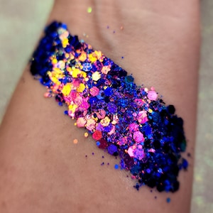 May include: A close-up of iridescent glitter in shades of blue, purple, pink, and gold. The glitter is arranged in a gradient pattern, with the darkest blue at the bottom and the lightest gold at the top.