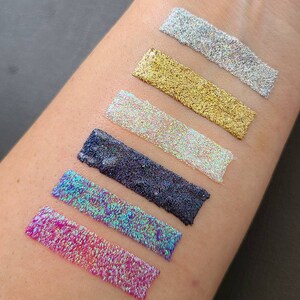 K&ouml;nnte beinhalten: Sechs verschiedene Farben von Glitter, darunter rosa, blau, schwarz, wei&szlig;, gold und silber, werden auf dem Arm einer Person gezeigt. Der Glitter ist in einer Linie angeordnet, wobei der rosa Glitter unten und der silberne Glitter oben ist.