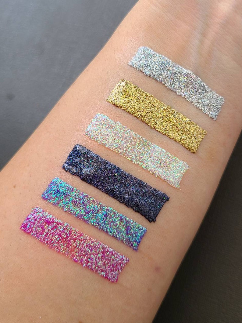 K&ouml;nnte beinhalten: Sechs verschiedene Farben von Glitter, darunter rosa, blau, schwarz, wei&szlig;, gold und silber, werden auf dem Arm einer Person gezeigt. Der Glitter ist in einer Linie angeordnet, wobei der rosa Glitter unten und der silberne Glitter oben ist.