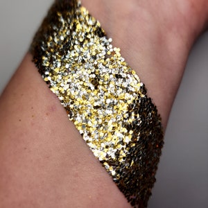 May include: A close-up of a person's arm with a diagonal stripe of gold and silver glitter applied to the skin.