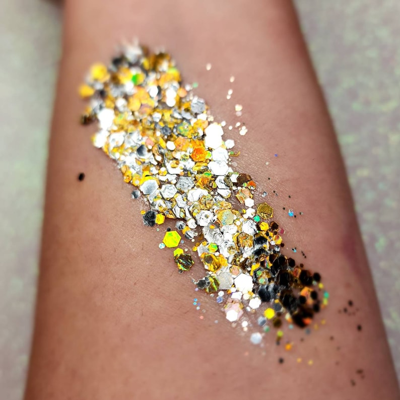 Puede incluir: Primer plano de un brazo de tono piel con una muestra de purpurina dorada, plateada y negra. La purpurina est&aacute; en una l&iacute;nea diagonal en el brazo.
