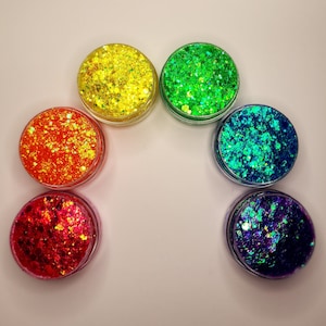Puede incluir: Siete pequeños frascos de purpurina iridiscente en varios colores, incluyendo amarillo, verde, naranja, rojo, azul y morado. Los frascos están dispuestos en un semicírculo.