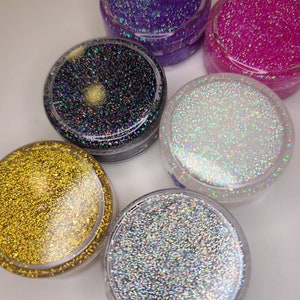 K&ouml;nnte beinhalten: Sechs kleine Gl&auml;ser mit Glitter, jedes gef&uuml;llt mit einer anderen Farbe von Glitter. Die Farben sind lila, rosa, schwarz, wei&szlig;, gold und silber.