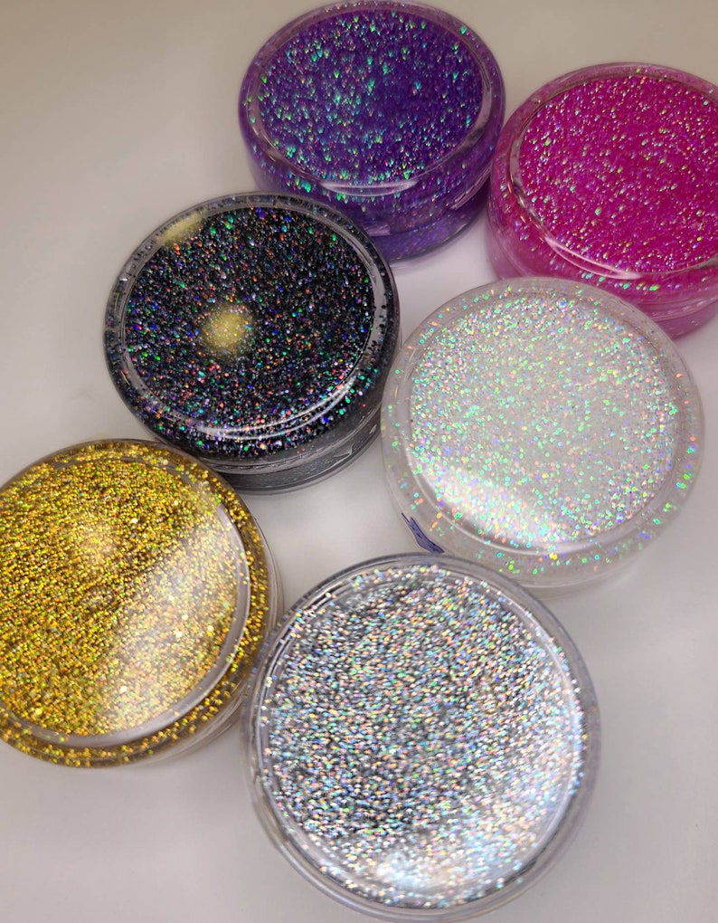 K&ouml;nnte beinhalten: Sechs kleine Gl&auml;ser mit Glitter, jedes gef&uuml;llt mit einer anderen Farbe von Glitter. Die Farben sind lila, rosa, schwarz, wei&szlig;, gold und silber.
