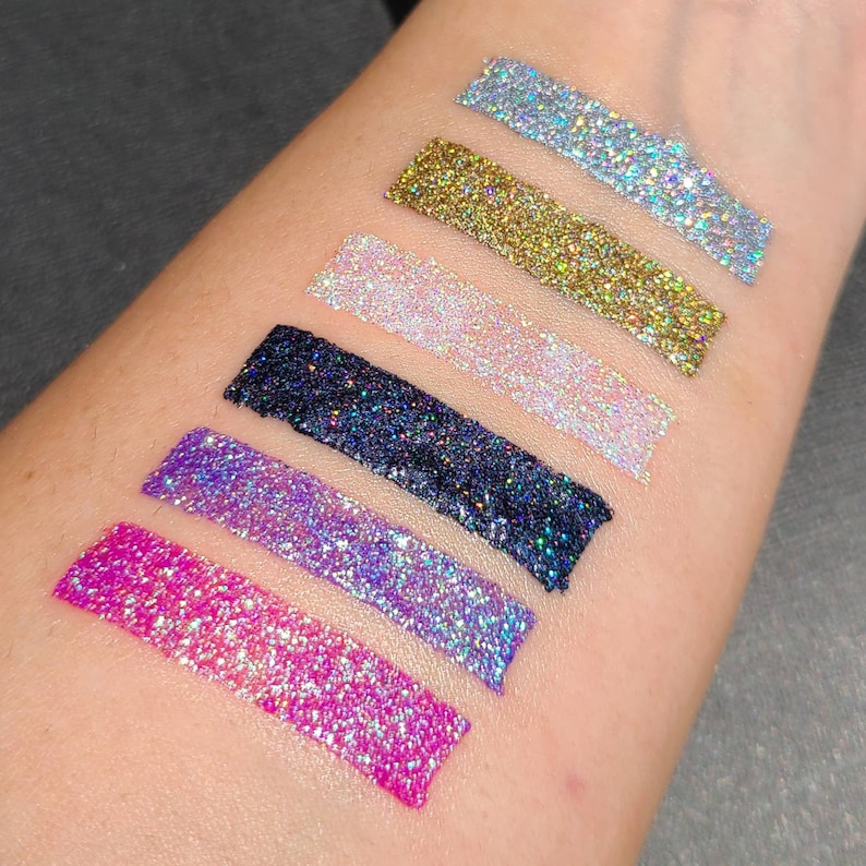 K&ouml;nnte beinhalten: Nahaufnahme von sechs verschiedenen Glitterfarben, darunter Pink, Lila, Blau, Schwarz, Wei&szlig; und Gold. Der Glitter wird in einer Linie auf den Arm einer Person aufgetragen.