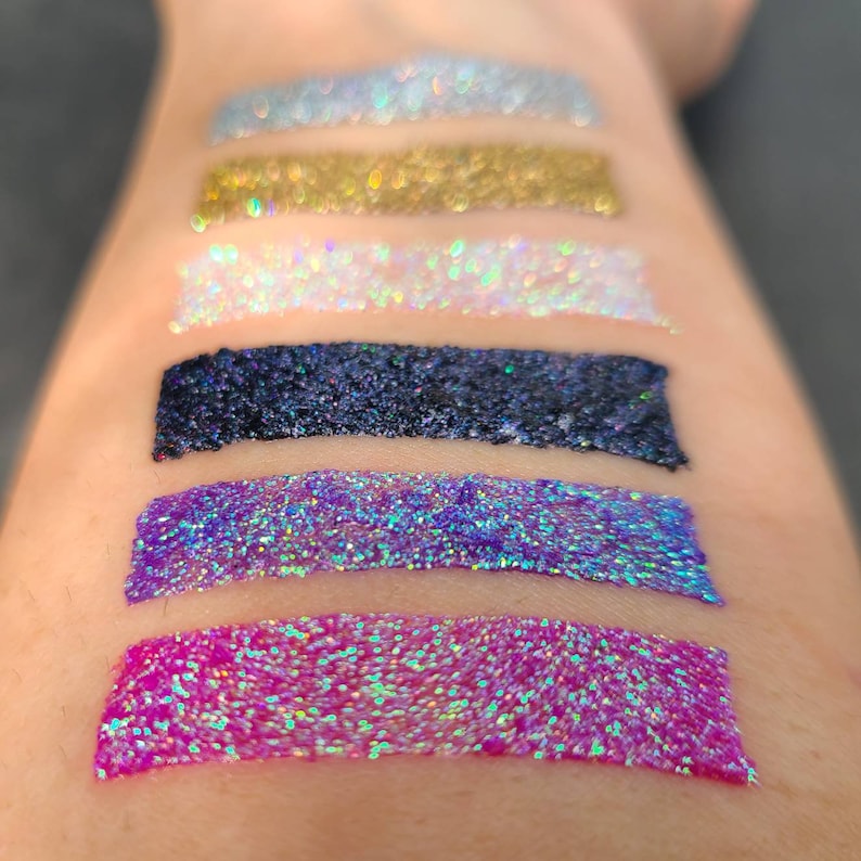 K&ouml;nnte beinhalten: F&uuml;nf Glitzerproben auf dem Arm einer Person. Die Proben sind in folgender Reihenfolge von oben nach unten angeordnet: hellblau, gold, wei&szlig;, schwarz, lila und rosa.