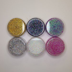 K&ouml;nnte beinhalten: Sechs kleine runde Gl&auml;ser gef&uuml;llt mit verschiedenen Farben von Glitter. Die Gl&auml;ser sind aus klarem Kunststoff und der Glitter ist durch den Kunststoff sichtbar. Die Farben des Glitters sind Gold, Blau, Lila, Silber, Wei&szlig; und Pink.