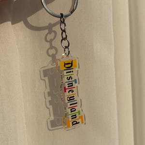 May include: Clear acrylic keychain with the word "Disneyland" in colorful letters. The keychain is attached to a silver chain and ring. The background is a light-colored fabric.