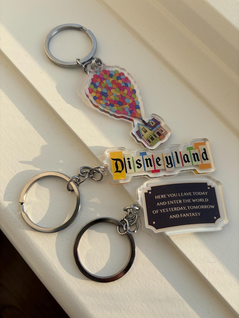 May include: Three keychains with silver rings. One keychain features a colorful balloon cluster and a house. Another says "Disneyland" in colorful letters. The third keychain has the text "HERE YOU LEAVE TODAY AND ENTER THE WORLD OF YESTERDAY, TOMORROW AND FANTASY."