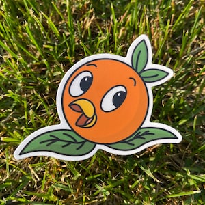 Puede incluir: Una pegatina troquelada con un personaje de dibujos animados de color naranja con una expresión alegre. El personaje naranja tiene ojos grandes y un pico amarillo, sobre hojas verdes. La pegatina está colocada sobre hierba verde.