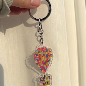 May include: A clear acrylic keyring featuring a colourful illustration of a house attached to a cluster of balloons. The balloons are in various colours, and the house has a yellow and brown facade. The keyring is attached to a silver ring.