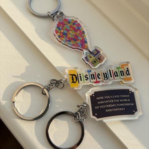 May include: Three keychains with silver rings. One keychain features a colorful balloon cluster and a house. Another says "Disneyland" in colorful letters. The third keychain has the text "HERE YOU LEAVE TODAY AND ENTER THE WORLD OF YESTERDAY, TOMORROW AND FANTASY."