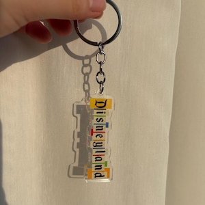 May include: Clear acrylic Disneyland keychain with a silver keyring and chain. The keychain features the word "Disneyland" in colorful, stacked letters. The background is a neutral color.