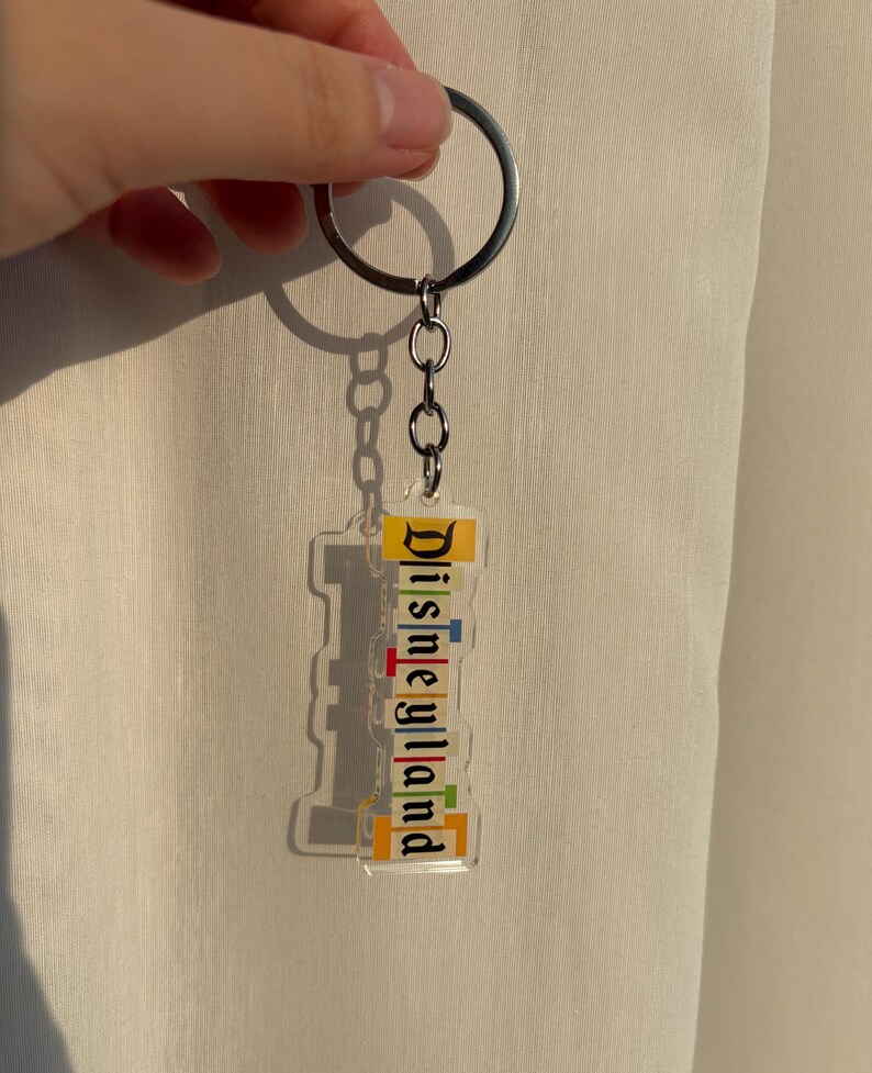 May include: Clear acrylic Disneyland keychain with a silver keyring and chain. The keychain features the word "Disneyland" in colorful, stacked letters. The background is a neutral color.