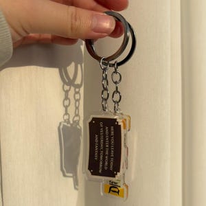 May include: A keychain with a silver ring and chain holding two rectangular acrylic charms. The charms have a brown border and text that reads "HERE YOU LEAVE TODAY AND ENTER THE WORLD OF YESTERDAY, TOMORROW AND FANTASY".