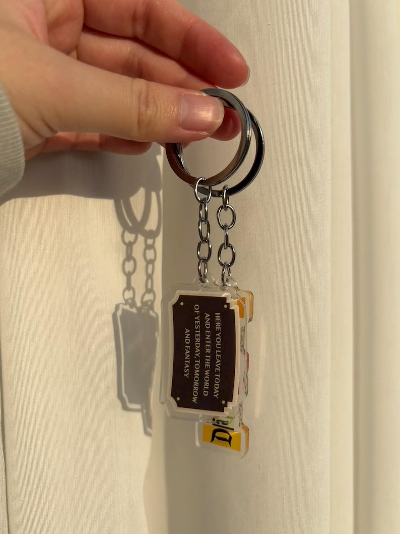 May include: A keychain with a silver ring and chain holding two rectangular acrylic charms. The charms have a brown border and text that reads "HERE YOU LEAVE TODAY AND ENTER THE WORLD OF YESTERDAY, TOMORROW AND FANTASY".