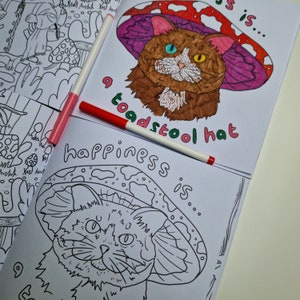 Op de afbeelding: Een kleurplaat met een zwart-witte illustratie van een kat die een paddenstoelhoed draagt. De tekst "happiness is... a toadstool hat" staat onder de kat.