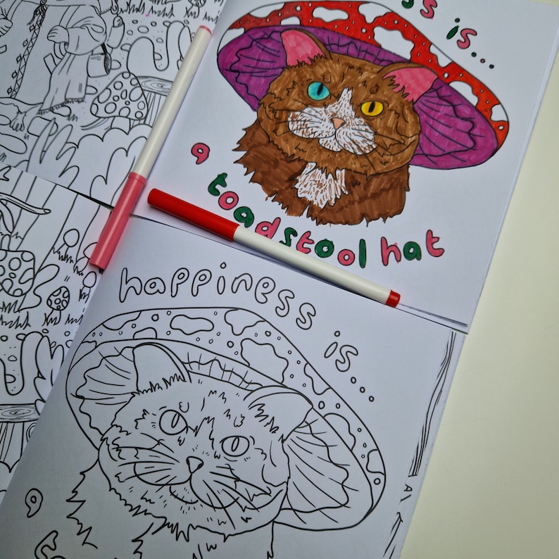 Op de afbeelding: Een kleurplaat met een zwart-witte illustratie van een kat die een paddenstoelhoed draagt. De tekst "happiness is... a toadstool hat" staat onder de kat.