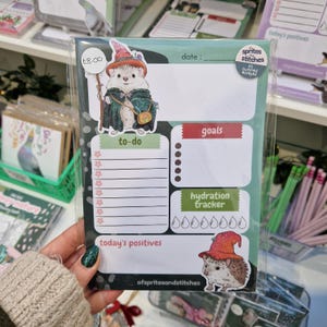 May include: A notepad featuring a hedgehog dressed as a wizard, with sections for to-do lists, goals, and a hydration tracker. The notepad is packaged in clear plastic.