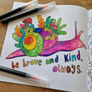 Op de afbeelding: Een kleurrijke illustratie van een slak met een regenboog-schelp en een roze lichaam. De slak is omringd door paddenstoelen en bloemen. De tekst "be brave and kind, always." staat onder de slak.