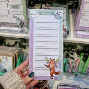 May include: A lined notepad with the text "take note" at the top. A cartoon fox with fairy wings is on the bottom right. The notepad is in a clear plastic sleeve. Other stationery items are visible in the background.