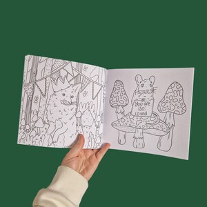 Op de afbeelding: Een open kleurboek met twee pagina's met zwart-wit illustraties. De linkerpagina toont een vos met een kroon in een bossetting met vlaggen en bomen. De rechterpagina toont een muis die een bord vasthoudt met de tekst "You are so loved" en op een paddenstoel zit.