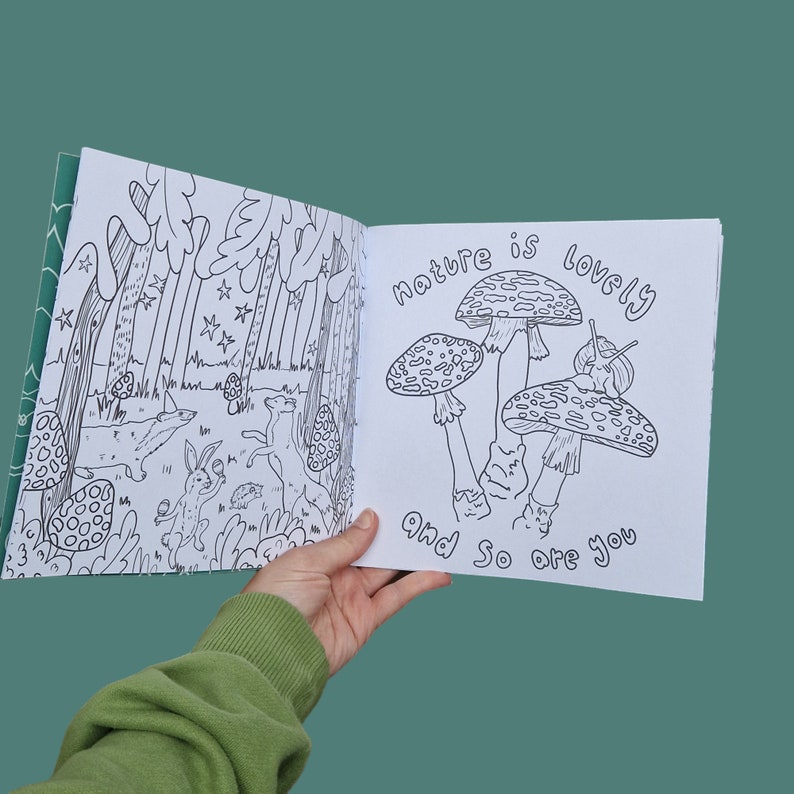 Op de afbeelding: Een open kleurboek met een zwart-witte illustratie van paddenstoelen en een bos sc&egrave;ne. De tekst op de pagina luidt "Nature is lovely and so are you."