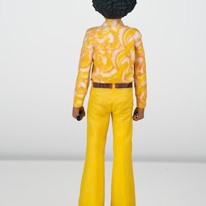 Puede incluir: Una figurita de una persona con un peinado afro que lleva una camisa de estampado amarillo y blanco, un cintur&oacute;n marr&oacute;n y pantalones acampanados amarillos.