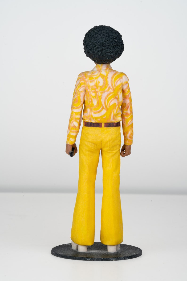 Puede incluir: Una figurita de una persona con un peinado afro que lleva una camisa de estampado amarillo y blanco, un cintur&oacute;n marr&oacute;n y pantalones acampanados amarillos.
