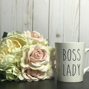 May include: White ceramic mug with black text that reads "BOSS LADY" on a white background. The mug is sitting on a black surface next to a bouquet of white and pink roses.