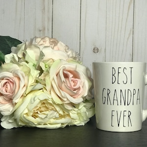 May include: A white ceramic mug with the text "BEST GRANDPA EVER" in black lettering. The mug is next to a bouquet of light pink and cream-colored roses and hydrangeas. The background is a light wood panel.