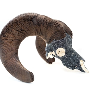 May include: A bighorn sheep skull with intricate black and silver designs on the bone. The horns are brown and have a rough, textured surface.