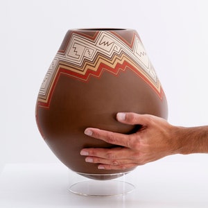 Puede incluir: Una olla de cerámica marrón con un diseño geométrico en rojo, blanco y marrón. La olla está sobre un soporte transparente.