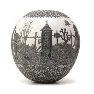 May include: A white ceramic sphere with a black and white design depicting a graveyard scene with skeletons, crosses, and a figure of the Virgin Mary. The text "COMUNION GENERAL QUEZADA" is visible on the sphere.