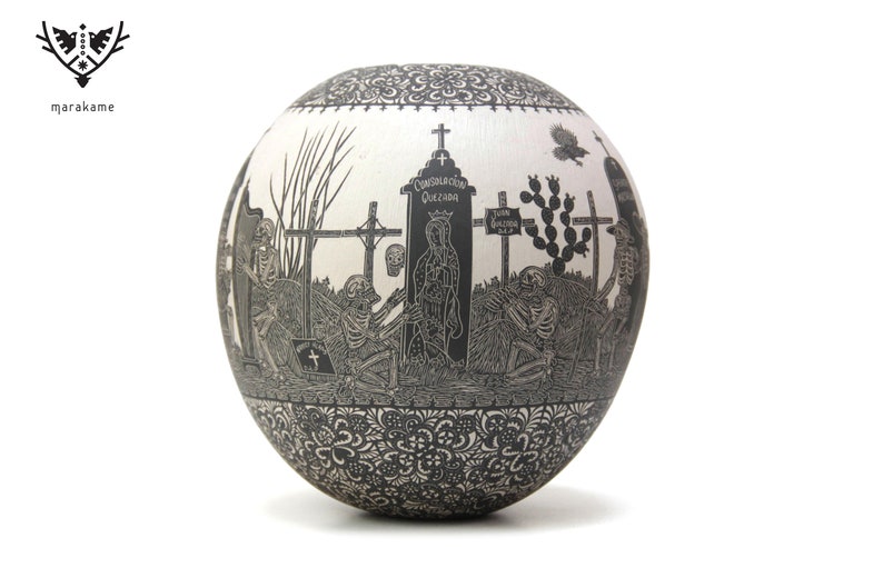 May include: A white ceramic sphere with a black and white design depicting a graveyard scene with skeletons, crosses, and a figure of the Virgin Mary. The text "COMUNION GENERAL QUEZADA" is visible on the sphere.